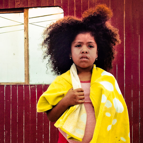 A young girl with afro hairstyle stands in front of a wooden house in Pachacútec, in the northern outskirts of Lima, Peru.
