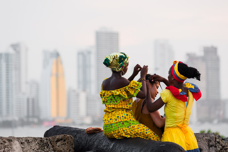 Afro-Colombian girls, dressed in the traditional ‘palenquera’ costume, create a braided hairstyle for a female tourist on the stone walls in Cartagena, Colombia.