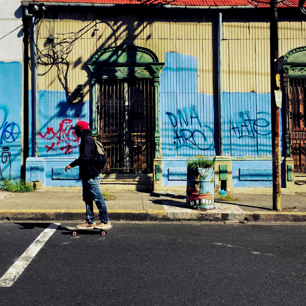 A Salvadoran young man rides a skateboard in front of a common lower middle class house, with Spanish colonial architecture elements and painted over by graffiti tags, built in the center of San Salvador, El Salvador.