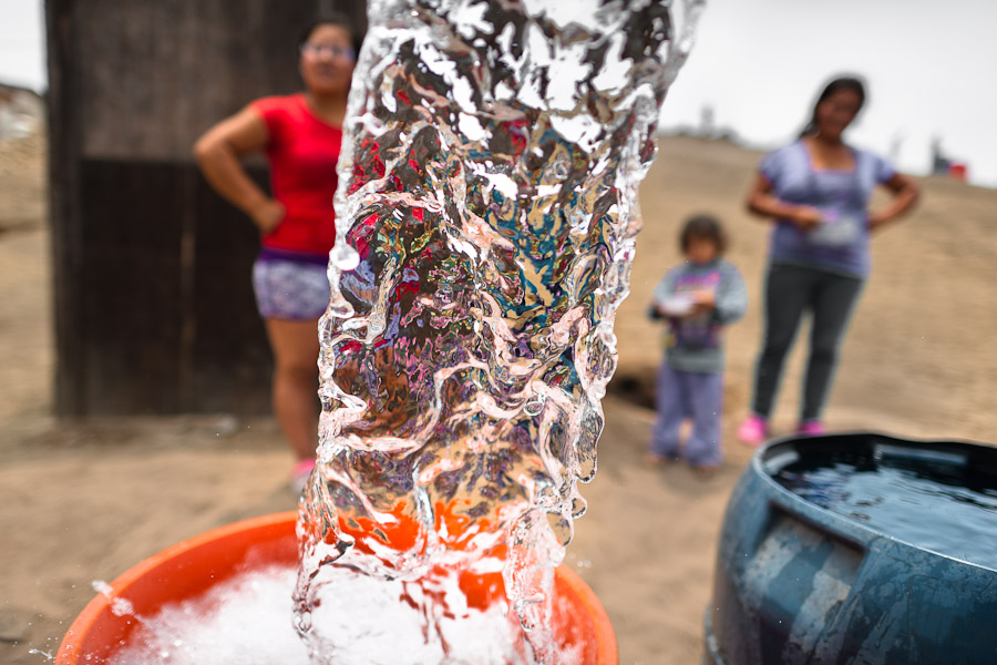 A splashing stream of drinking water falls into a plastic barrel in front of a wooden house on the dusty hillside of Pachacútec, a desert suburb of Lima, Peru.