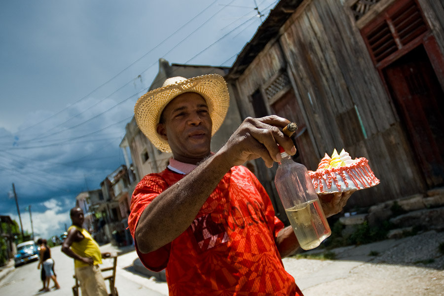 A Cuban man hurries for a fiesta, holding a bottle of rum, smoking a cigar and carrying a sweet cake. Although the overall situation in Cuba is tough, Cubans never forget to celebrate, to call for a fiesta.