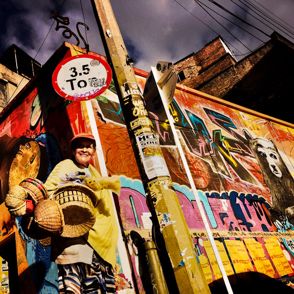 A Colombian street vendor walks around a street corner, covered in colorful graffiti artwork and posters, in La Candelaria, Bogota, Colombia.
