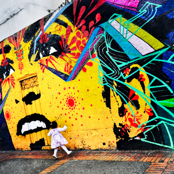 A Colombian baby girl jumps in front of a graffiti artwork, created by artists named Stinkfish & Zas, in La Candelaria, Bogotá, Colombia.