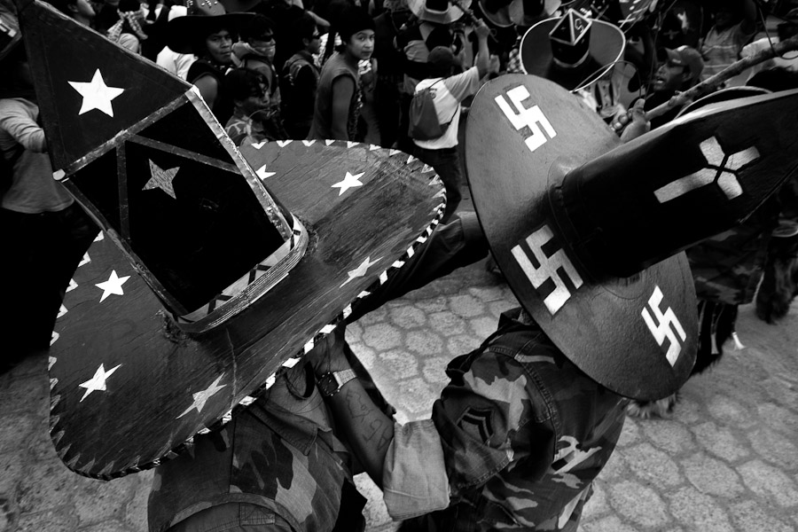 Indian dancers wear tall, broad-rimmed, black cardboard hats, painted over by religious and political symbols, like a Christian cross, a swastika or a star.