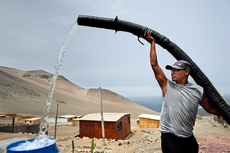 A Peruvian water distribution worker with a hose splashes drinking water into a plastic barrel on the dusty hillside of Pachacútec, a desert suburb of Lima, Peru.