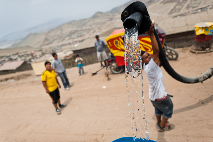 A Peruvian water distribution worker with a hose splashes drinking water from a truck into a plastic barrel on the dusty hillside of Pachacútec, a desert suburb of Lima, Peru.