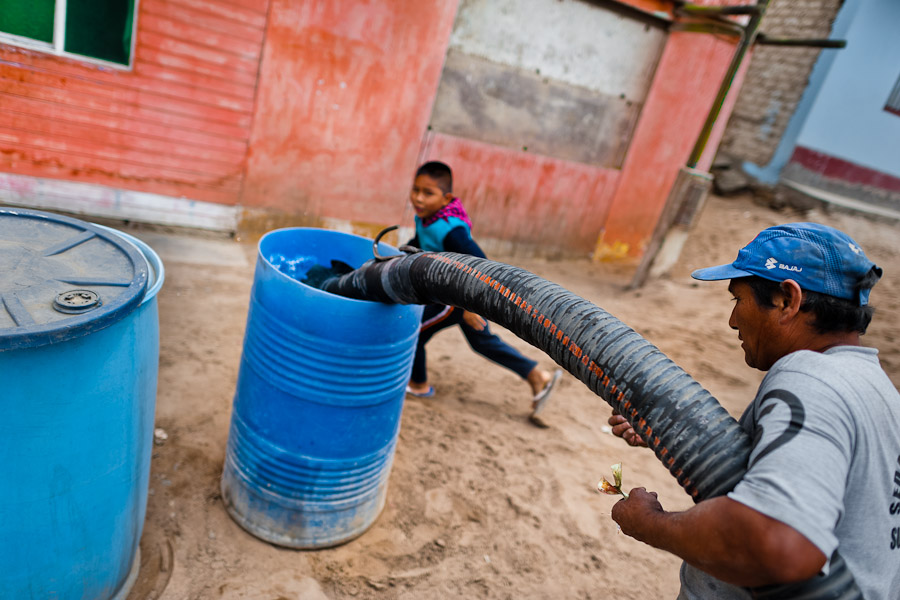 A Peruvian water distribution worker with a pipe fills a plastic barrel with drinking water on the dusty hillside of Pachacútec, a desert suburb of Lima, Peru.